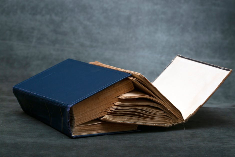 A worn-out vintage book with blue hardcover resting open on a dark surface, displaying aged pages.